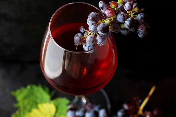 Ein Weinglas mit rotem Wein, darauf liegend eine Traube, umgeben von grünen Blättern.