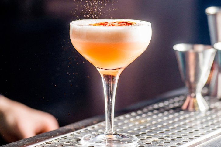 Ein Cocktailglas mit Schaum, garniert, steht auf einer metallischen Theke, umgeben von Barzubehör.