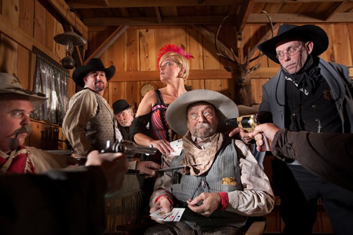 Personen sitzen mit Pistolen in einem rustikalen Raum, einige halten Spielkarten.