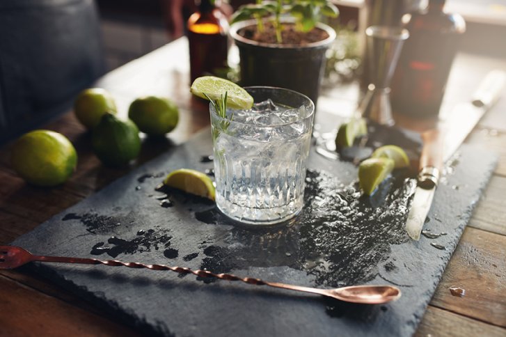 Ein Glas Gin Tonic mit Limettenscheiben steht auf einem nassen Schieferbrett zwischen Limetten.
