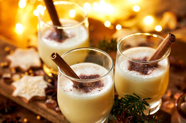 Gläser mit Eierlikör und Zimtstangen auf Holztisch, umgeben von weihnachtlichen Keksen und Lichtern.