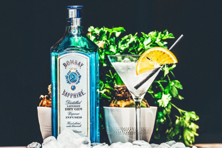 Blaue Ginflasche steht neben Cocktailglas, dekoriert mit Orange, umgeben von Eis und Pflanzen.