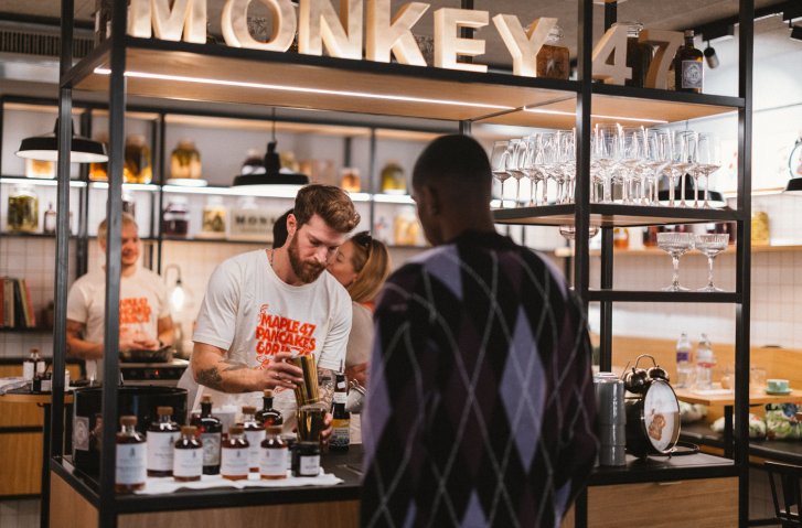 Barmann mixt Getränke, umgeben von Flaschen, Gläsern und Regalen, unter dem "MONKEY 47"-Schild.