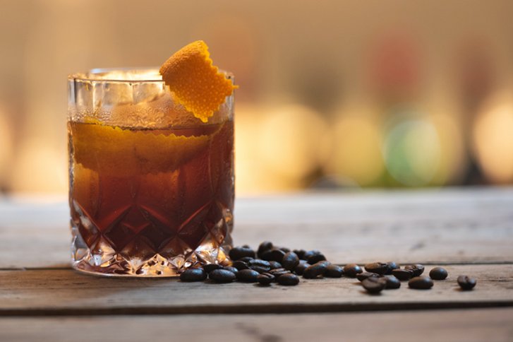 Ein Cocktailglas mit Eiswürfeln und Orangenschale steht auf Holztisch, umgeben von Kaffeebohnen.
