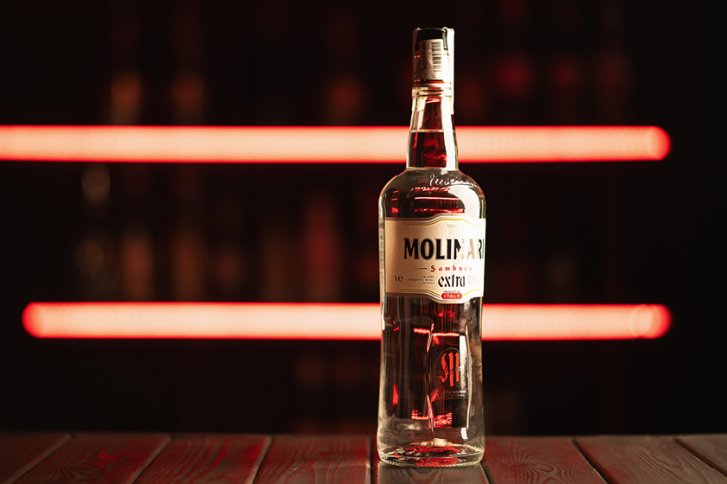 Flasche Molinari Sambuca steht auf Holztisch vor unscharfem Hintergrund mit roten Lichtern.