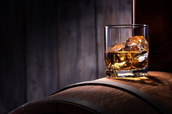 Glas mit Eiswürfeln und Whisky, stehend auf einem Holzfass, vor dunklem Hintergrund.