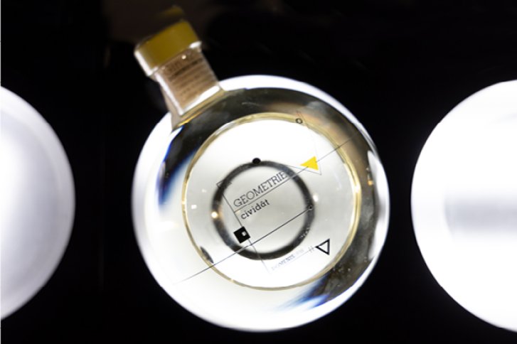 Eine Flasche von oben, gefüllt mit klarer Flüssigkeit, auf schwarzem Hintergrund. Text: GEOMETRIE.