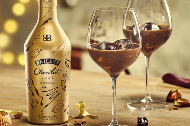 Eine goldene Baileys-Flasche steht neben zwei Gläsern mit Eis, auf einem Holztisch.