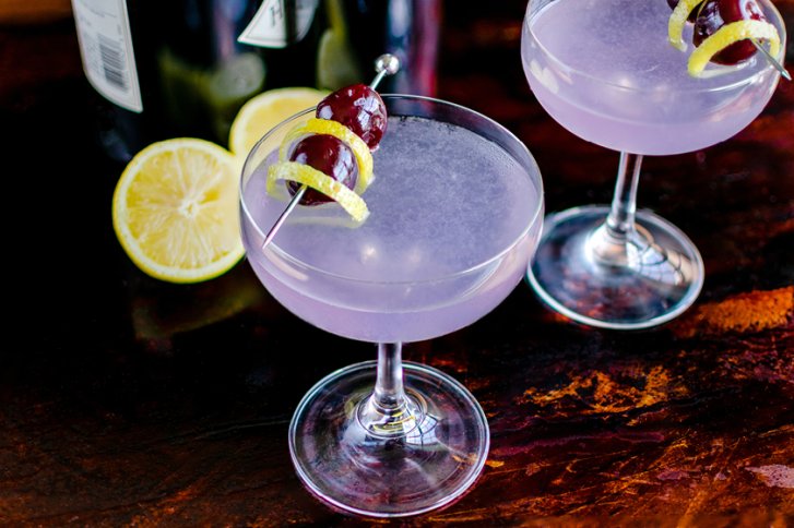 Zwei lila Cocktails, dekoriert mit Zitronenschale und Kirschen, stehen auf dunklem Holztisch.