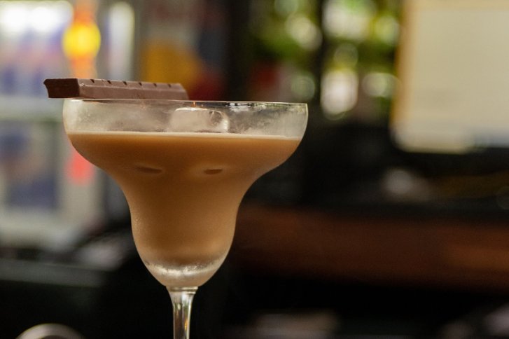Ein Cocktailglas mit braunem Getränk, dekoriert mit Schokoladenstück, steht in einer Barumgebung.