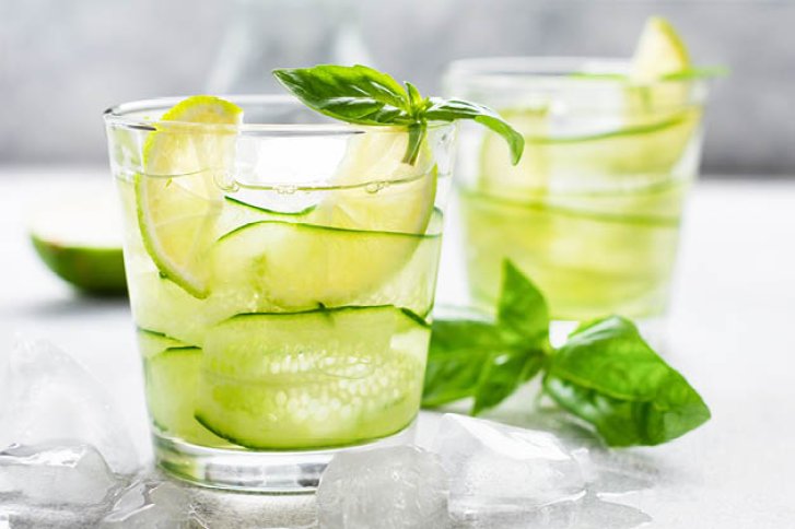 Glas mit Gurkenscheiben und Limette, dekoriert mit Basilikum, umgeben von Eiswürfeln auf Tisch.