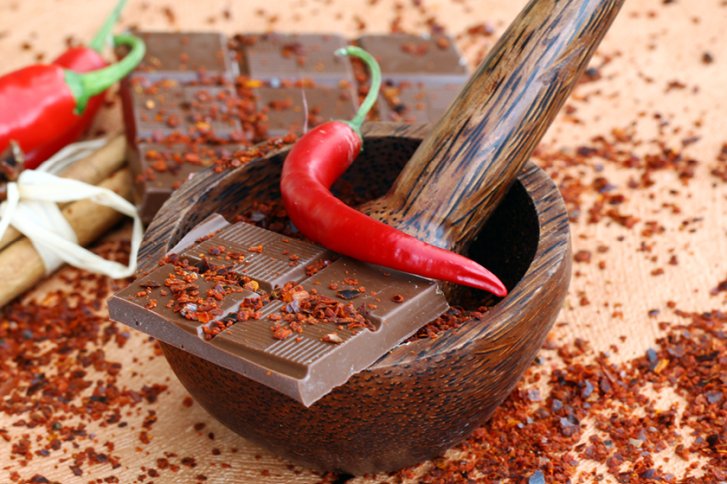 Schokolade und Chili liegen in einer Holzschale mit Mörser, umgeben von Chiliflocken.