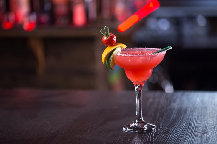 Roter Cocktail im Glas, garniert mit Erdbeere und Limette, steht auf dunklem Tresen.