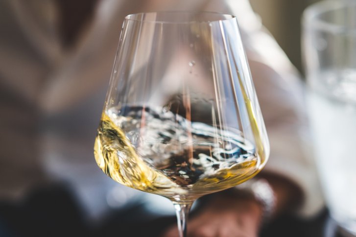 Weinglas mit Weißwein, schwenkt in einer Person Hand, unscharfer Hintergrund.