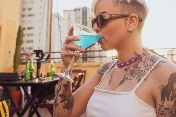 Person trinkt blauen Cocktail auf sonniger Terrasse mit sichtbaren Tätowierungen und Sonnenbrille.
