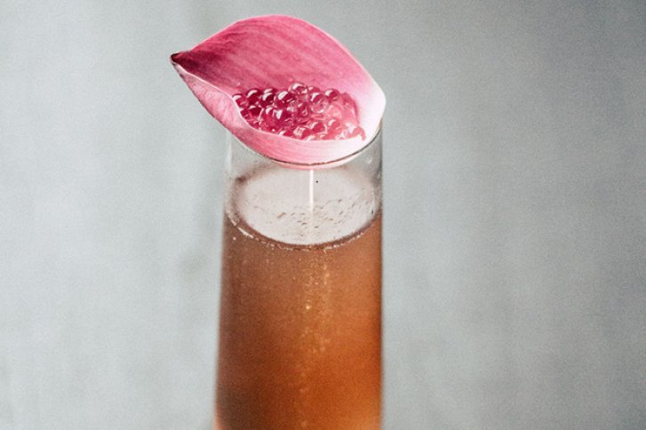 Ein Glas brauner Cocktail, dekoriert mit rosa Blütenblatt und Perlen, steht auf grauem Untergrund.