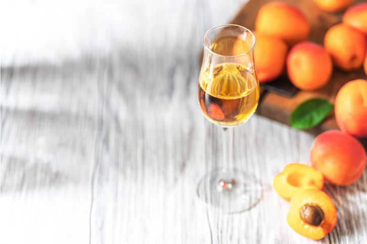 Ein Glas Schnaps steht auf hellem Holztisch, umgeben von frischen Aprikosen und einem Holzbrett.
