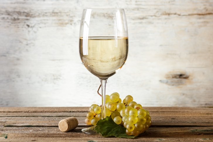 Weinglas steht auf Holztisch neben weißen Trauben und Korken, hellgraue Wand im Hintergrund.