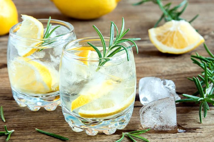 Zwei Gläser mit Zitronen und Rosmarin, stehen auf Holztisch, umgeben von Eiswürfeln und Zitronenstücken.