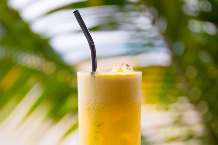 Gelbes Getränk mit Schaum und Strohalm, eingefangen vor verschwommenen grünen Blättern im Hintergrund.