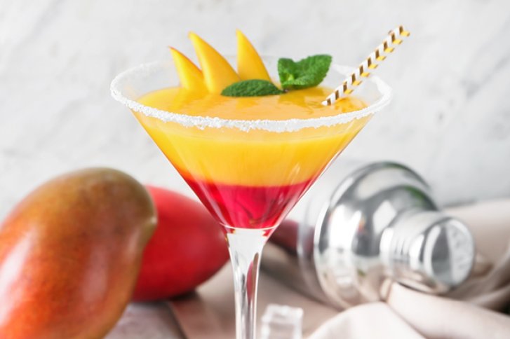 Ein Cocktailglas mit gelb-rotem Getränk, Mangoscheiben und Minze, steht neben einem Shaker.