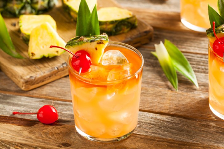 Ein Cocktailglas mit Eis, Ananasstück, Kirsche; auf Holztisch mit ganzen Ananasstücken.