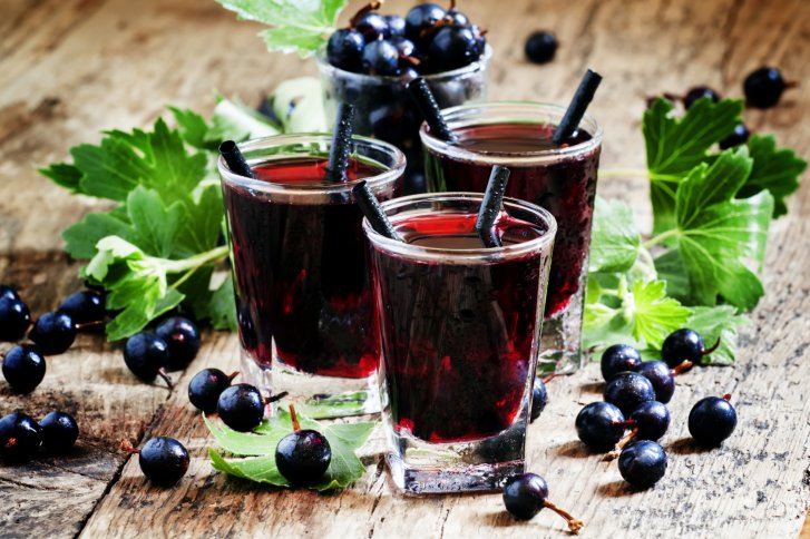 Drei Gläser mit dunklem Beerensaft stehen auf einem Holztisch, umgeben von schwarzen Johannisbeeren.