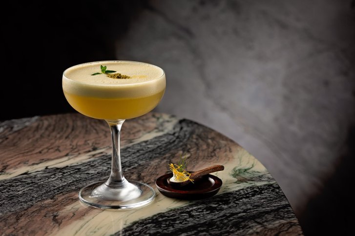 Cocktailglas steht auf marmoriertem Tisch, dekoriert mit Minze; Löffel daneben. Düstere Umgebung.