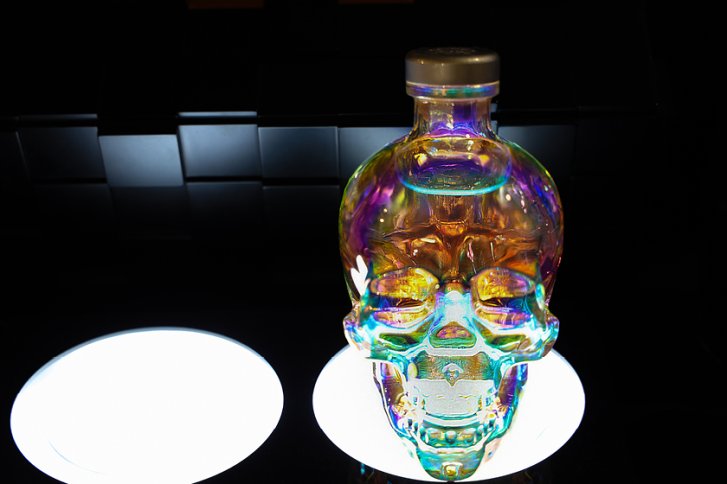 Eine schädelähnliche, regenbogenfarbene Flasche steht beleuchtet vor einem dunklen, gefliesten Hintergrund.