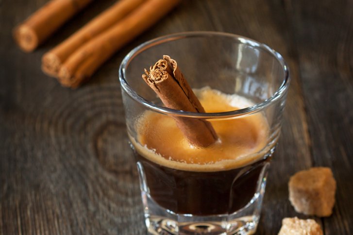 Ein Glas Kaffee mit Zimtstange steht auf Holztisch, umgeben von Zuckerwürfeln.