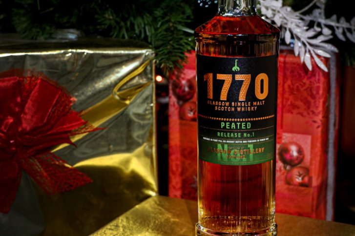 Eine Whiskyflasche steht dekorativ zwischen weihnachtlich verpackten Geschenken und Tannenzweigen.