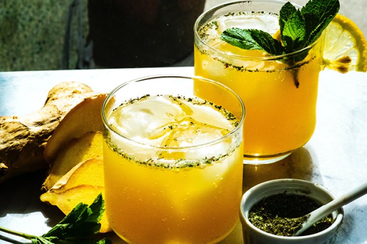 Zwei gelbe Cocktails mit Eis und Minze stehen auf Tisch neben Ingwer und Gewürzschale.