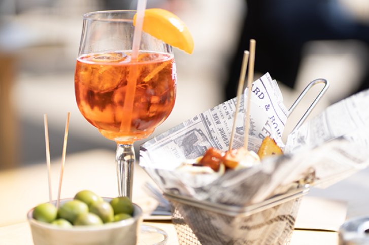 Ein Aperol-Glas mit Orangenscheibe steht neben Oliven und Snacks auf einem Tisch draußen.