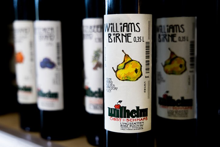 Flaschen stehen nebeneinander im Regal, etikettiert mit „Williams Birne“ und Fruchtillustrationen.