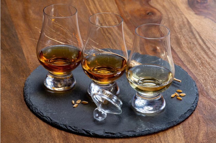 Drei Whisky-Gläser stehen auf einer Schieferplatte, umgeben von Weizenkörnern auf Holztisch.