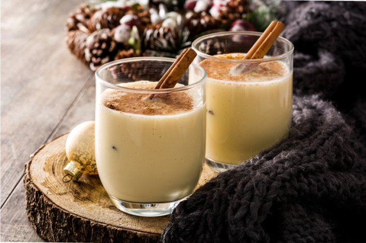 Zwei Gläser Eierlikör stehen auf Holzteller, dekoriert mit Zimtstangen und Weihnachtskugeln.