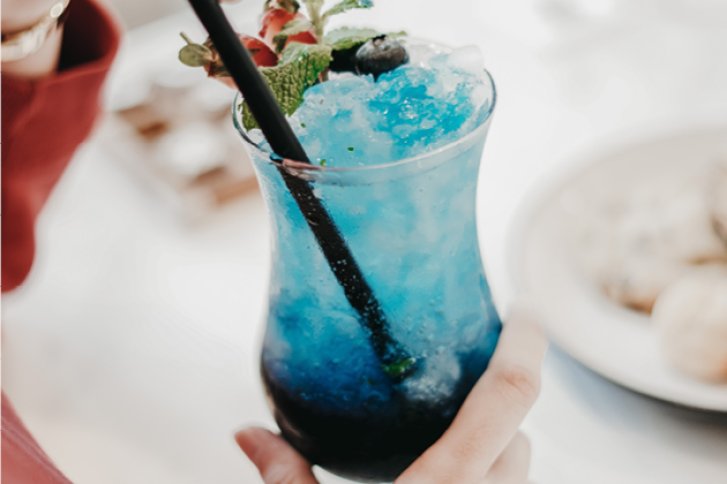 Ein blaues Cocktailglas wird von Hand gehalten, daneben ein Teller auf hellen Tisch.
