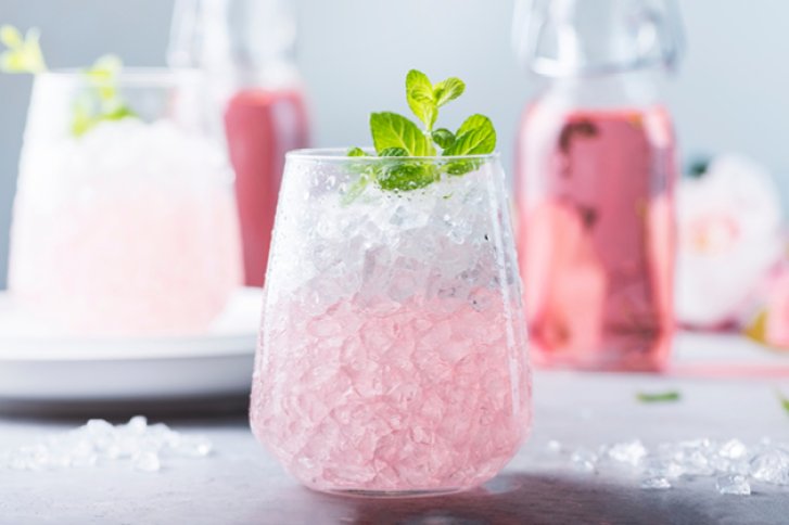 Ein Glas mit rosa Getränk, gefüllt mit Eis und Minze, auf grauem Hintergrund.