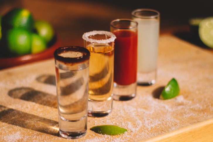 Vier bunte Shotgläser stehen auf Holzbrett mit Limetten und Salz umgeben.