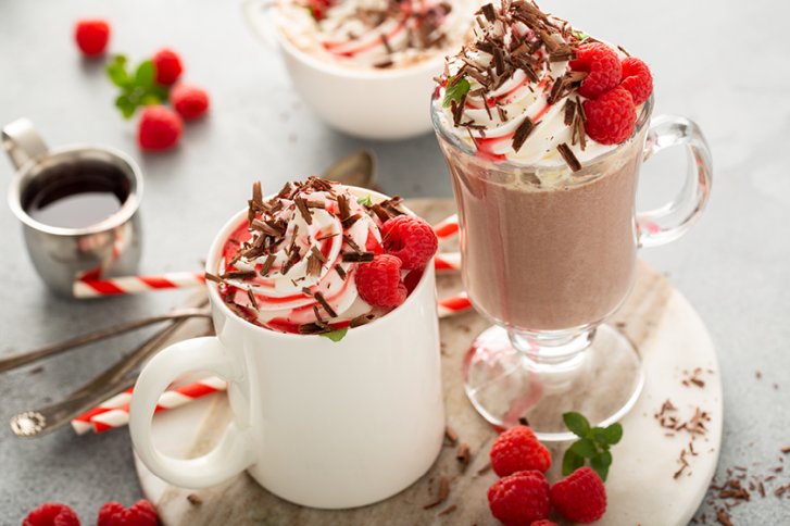 Heiße Schokoladen mit Sahne, Schokoraspeln und Himbeeren dekoriert, stehen auf einem Tisch mit Minze.