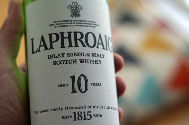 Flasche Whisky wird von Hand gehalten, Etikett mit "Laphroaig 10 Years" erkennbar, bunte Hintergründe.