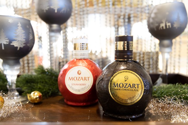 Zwei Mozart-Likörflaschen stehen dekorativ auf Holztisch, umgeben von weihnachtlichen Verzierungen und Gläsern.