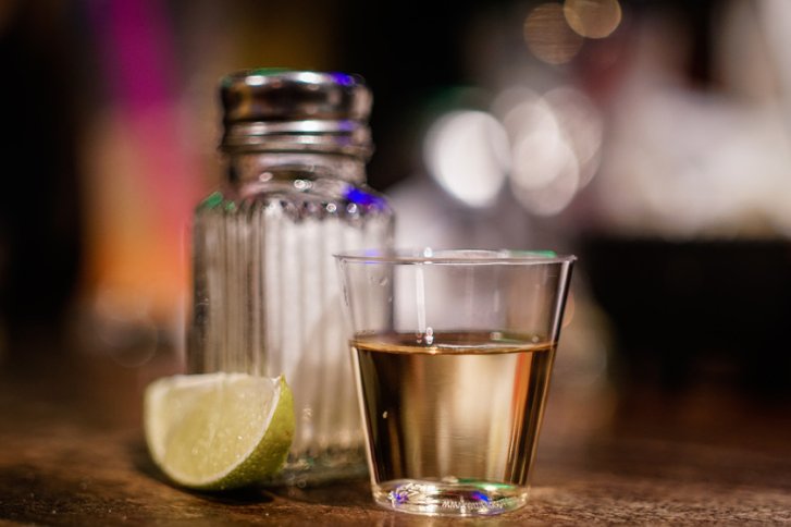 Ein Plastikglas mit Tequila steht neben Limette und Salzstreuer auf einem Holztisch.