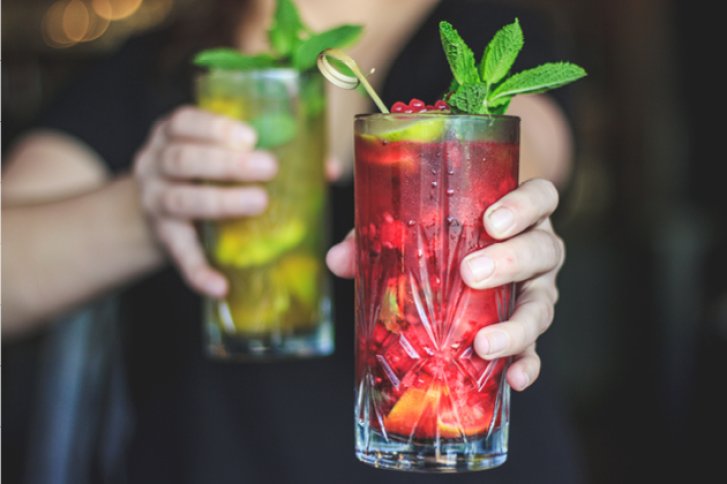 Zwei Gläser mit bunten Cocktails werden von einer Person mit grüner Garnitur gehalten.