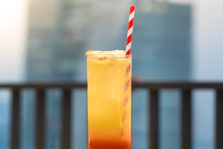 Ein Cocktail mit Schaum, rotem und weißem Strohhalm auf einer Terrasse vor Gebäuden.