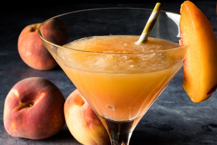 Ein Cocktailglas enthält ein pfirsichfarbenes Getränk mit Strohalm, umgeben von reifen Pfirsichen.