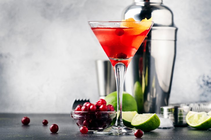Rotes Cocktail-Glas steht auf Tisch, umgeben von Limetten, Cocktailshaker und Preiselbeeren in stilvoller Umgebung.