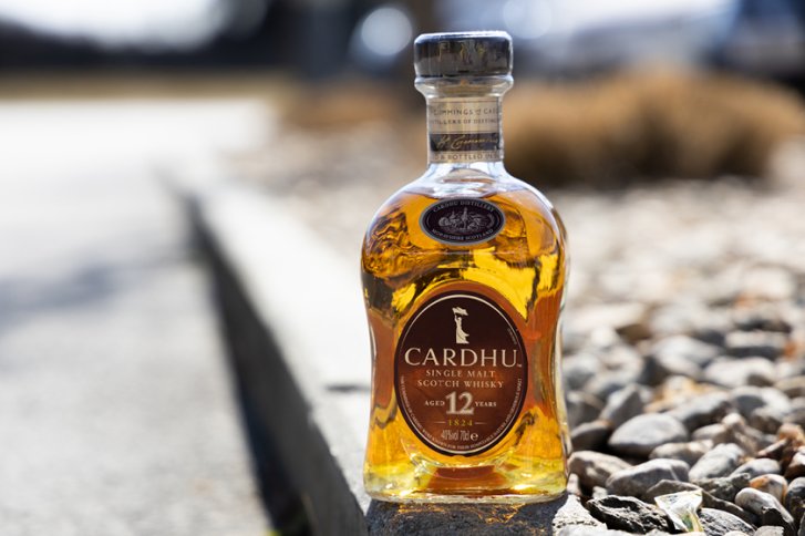 Flasche Cardhu Whisky steht auf Bordstein neben Kieselsteinen im Sonnenlicht.