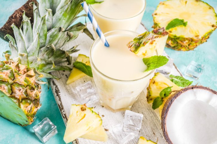 Piña Colada im Glas mit Strohhalm, Ananas und Kokos auf hellem Hintergrund.