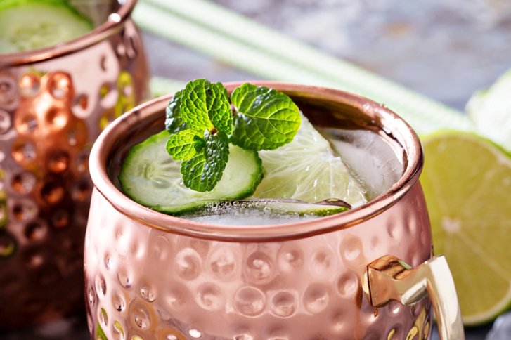 Kupferbecher mit Minze, Gurken- und Limettenscheiben gefüllt, auf einem Tisch mit hellgrünem Hintergrund.